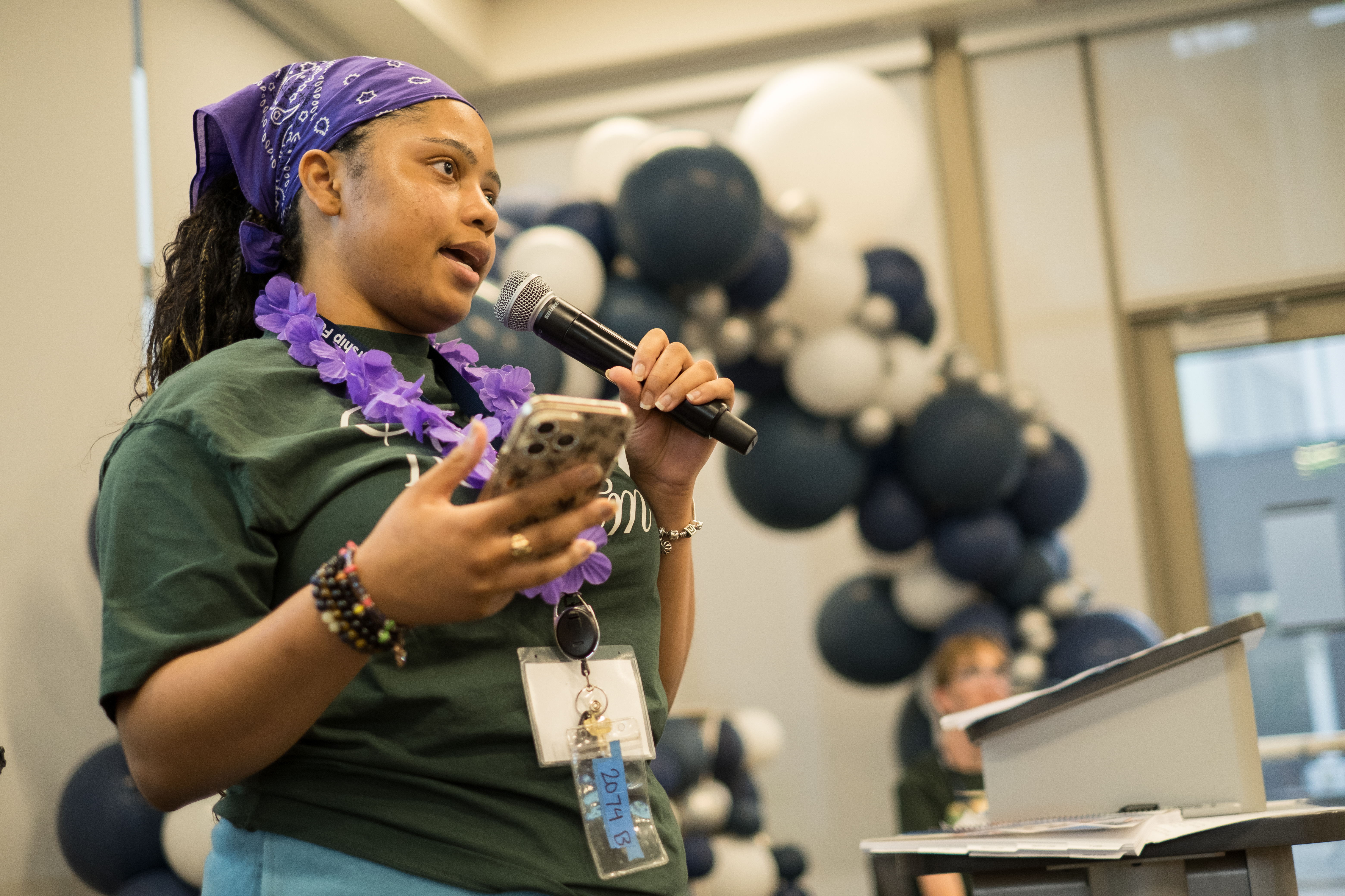 A person wearing a purple bandana and lei speaks into a microphone while holding a phone, standing in front of a large balloon display.